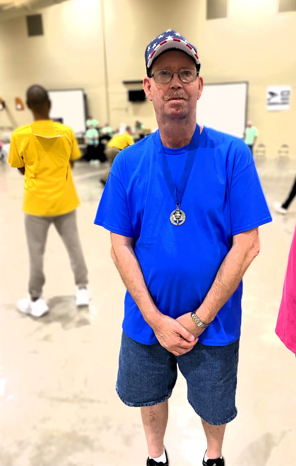 Adult male, standing, blue T-shirt and red, white and blue baseball cap with gold medal.