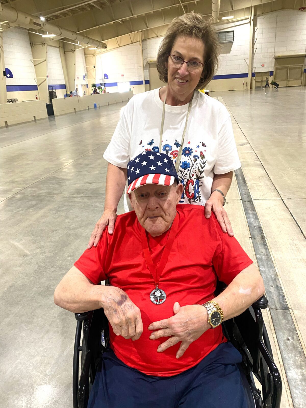 Adult senior male in wheelchair wearing red T-shirt with silver medal. Adult female white T-shirt standing behind him.
